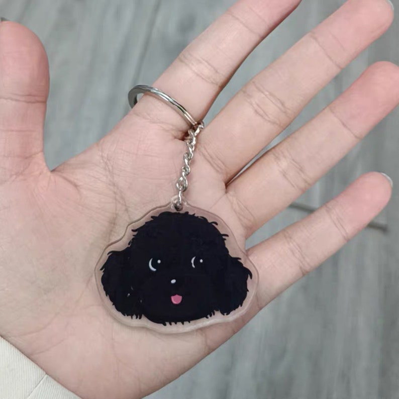 May include: A black and white cartoon poodle keyring with a pink tongue. The keyring is made of clear acrylic and has a silver metal ring.