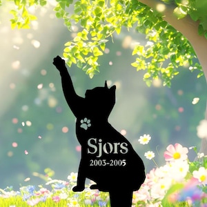 May include: A black cat silhouette garden stake featuring a paw print cutout, the name Sjors, and the years 2003-2005. The cat is depicted in a seated position with one front paw raised upward as if reaching toward the branches of a tree.