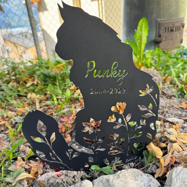 May include: A black metal silhouette of a cat with a long, curved tail, featuring cut-out floral patterns at the base. The name Punky and the years 2004-2023 are laser-cut into the center of the cat's body, creating a memorial garden stake.