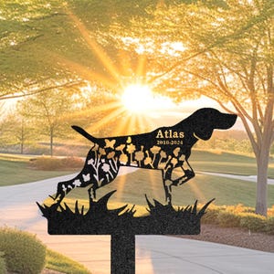 Può includere: Silhouette in metallo nero di un cane con dettagli floreali, in piedi su una base erbosa. Il nome del cane "Atlas" e le date "2010-2024" sono incise sulla silhouette.