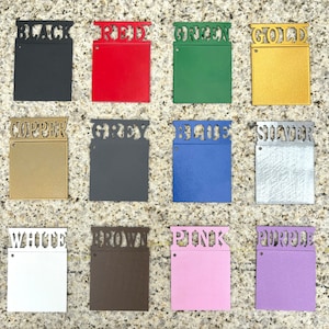 May include: A set of twelve square metal color swatches arranged in a grid. Each swatch features a cutout label at the top identifying the color: black, red, green, gold, copper, grey, blue, silver, white, brown, pink, and purple.