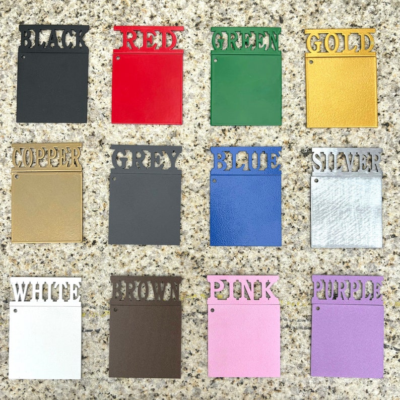 May include: A set of twelve square metal color swatches arranged in a grid. Each swatch features a cutout label at the top identifying the color: black, red, green, gold, copper, grey, blue, silver, white, brown, pink, and purple.