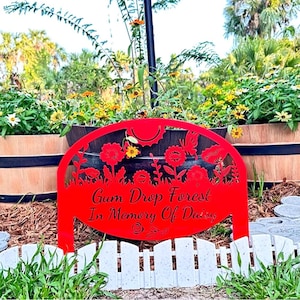 May include: A red metal garden sign with a semi-circular top featuring cutouts of flowers and a butterfly. The text reads Gum Drop Forest In Memory Of Daisy. The sign is positioned in front of a dark planter and a white picket fence border.