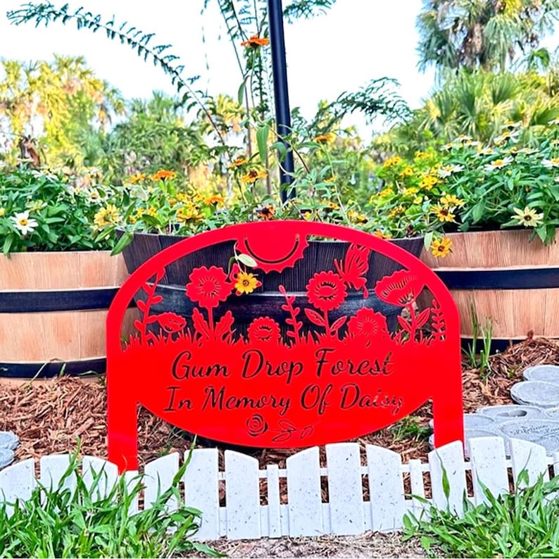 May include: A red metal garden sign with a semi-circular top featuring cutouts of flowers and a butterfly. The text reads Gum Drop Forest In Memory Of Daisy. The sign is positioned in front of a dark planter and a white picket fence border.
