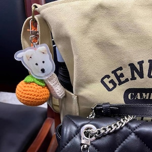 May include: A beige canvas tote bag with the text "GENZ CAMP" printed on it. A white dog keychain with a crocheted orange fruit charm is attached to the bag.