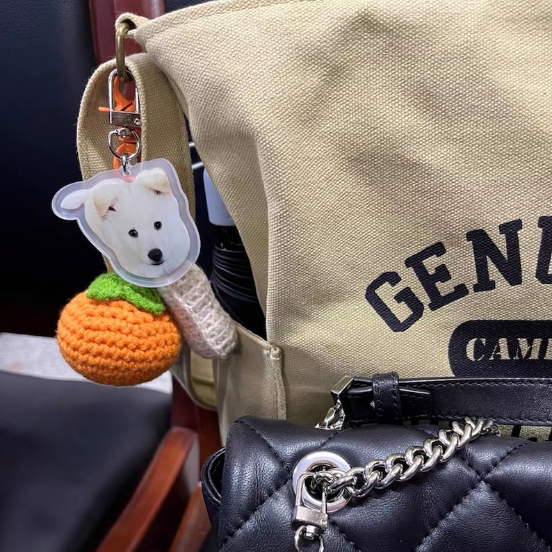 May include: A beige canvas tote bag with the text "GENZ CAMP" printed on it. A white dog keychain with a crocheted orange fruit charm is attached to the bag.