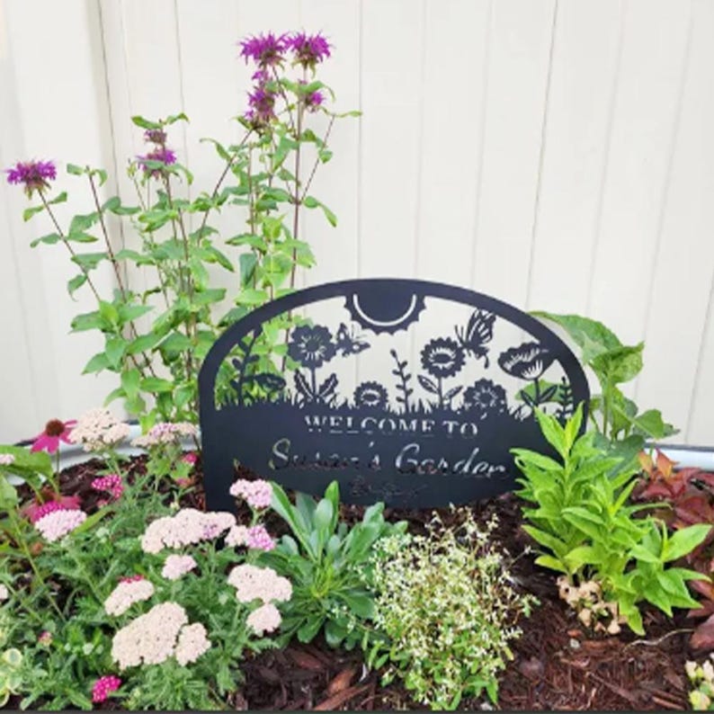 May include: A black metal garden sign with an arched top featuring cut-out designs of flowers, a sun, and a butterfly. The sign displays the text "WELCOME TO Susan's Garden" in a combination of block and script lettering, positioned among plants.