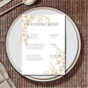 May include: A white wedding menu card with gold floral illustrations. The menu includes a Tuscan garden salad, wild-caught salmon, and white cake with almond flavor and lemon curd.