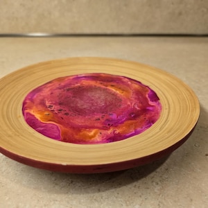 May include: A round wooden coaster with a pink, orange, and white resin inlay. The coaster has a natural wood finish and a dark red rim.