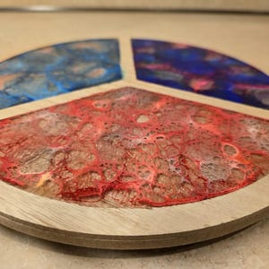 Puede incluir: Un posavasos de madera con tres secciones, cada una llena de resina de un color diferente. Las secciones son roja, azul y morada. La resina tiene un efecto marmoleado.