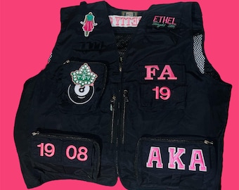 Op de afbeelding: Zwarte vest met meerdere zakken en ritsen. Voorzien van roze geborduurde tekst en patches, waaronder "FA 19", "AKA" en "19 08". Versierd met een jurk en een biljartbal patch. De vest heeft een mesh paneel aan de rechterkant.