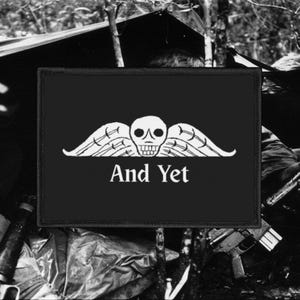 May include: Black rectangular patch with a white skull and wings design, and the text "And Yet". The patch is likely made of fabric and is designed to be sewn or ironed onto clothing or equipment. The design is stark and striking.