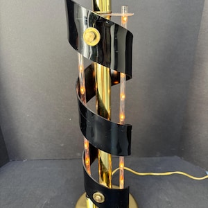 May include: A brass-colored table lamp with a spiral design. The lamp features a gold base and a central column with black, curved accents. Small lights are visible within the clear tubes. The lamp has a cord for power.