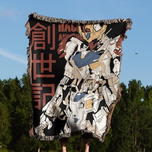 May include: A woven tapestry throw blanket featuring a black and white illustration of a robot with a yellow helmet. The robot is standing in a fighting pose. The text "HACKING AND VIN" is visible in the background. The blanket has a red fringe border and the Japanese characters "創造世界" are visible on the left side of the blanket.