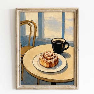 Könnte beinhalten: Ein gerahmter Kunstdruck zeigt eine Zimtschnecke mit weißem Zuckerguss auf einem weißen Teller, eine schwarze Kaffeetasse und einen Holzstuhl an einem runden Tisch. Die Szene befindet sich vor einem Fenster mit Blick auf einen blassblauen und gelben Himmel.