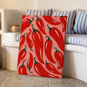 Könnte beinhalten: Ein rechteckiger Leinwanddruck mit einem sich wiederholenden Muster aus leuchtend roten Chilischoten mit grünen Stielen auf einem einfarbigen hellrosa Hintergrund. Das Kunstwerk zeigt mehrere Schoten in verschiedenen Richtungen.