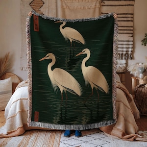 May include: A dark green woven throw blanket featuring three cream-colored herons standing in water. The design includes a vertical red rectangular stamp with black text in the upper left and a smaller red square stamp in the lower left corner.