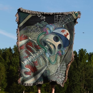 May include: A woven tapestry throw blanket featuring a skeleton wearing a blue and white hat with a number one on it. The skeleton is holding a green tube in its mouth and is surrounded by a web and bubbles.