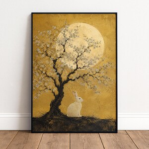 Könnte beinhalten: Ein gerahmter Kunstdruck, der einen weißen Hasen unter einem dunklen, knorrigen Baum mit weißen Blüten zeigt. Der Hintergrund ist in strukturiertem Goldgelb gehalten, mit einem großen, blassen, kreisförmigen Mond hinter den Ästen.