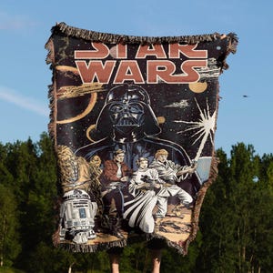 May include: A woven tapestry featuring characters from Star Wars, including Darth Vader, Luke Skywalker, Princess Leia, Chewbacca, and R2-D2. The tapestry is black with a dark blue background and features a white starburst in the upper right corner. The text "STAR WARS" is printed in red at the top of the tapestry.