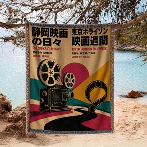 May include: A woven throw blanket featuring a vintage film projector and film strip graphic. The design includes text in Japanese and English, such as Shizuoka Film Days 1989 and Tokyo Horizon Film Week 2001, set against a colorful retro background.