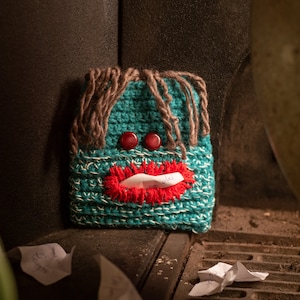 Può includere: Un copri scatola di fazzoletti in crochet color turchese con una bocca sorridente rossa e due occhi a bottone rossi. Il copri scatola è appoggiato su una superficie marrone con alcuni pezzi di carta bianca stropicciati.