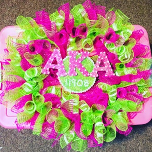 May include: A pink and green wreath with the letters "AKA" and the year "1908" in the center. The wreath is made of mesh and has a sparkly finish.