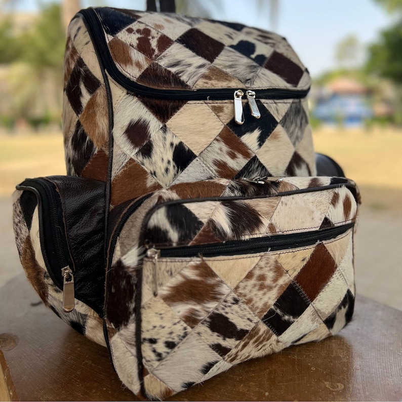May include: A patchwork backpack with a geometric pattern of brown, tan, black, and white cowhide. The backpack has a top flap, a front pocket, and two side pockets, all with zippered closures. The bag is designed for carrying items.