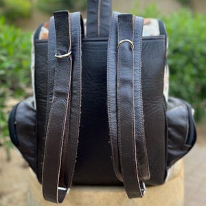 May include: Back view of a black leather backpack with brown leather straps and accents. The backpack features a patterned design on the sides and a zipper closure. The straps are attached with silver metal rings.