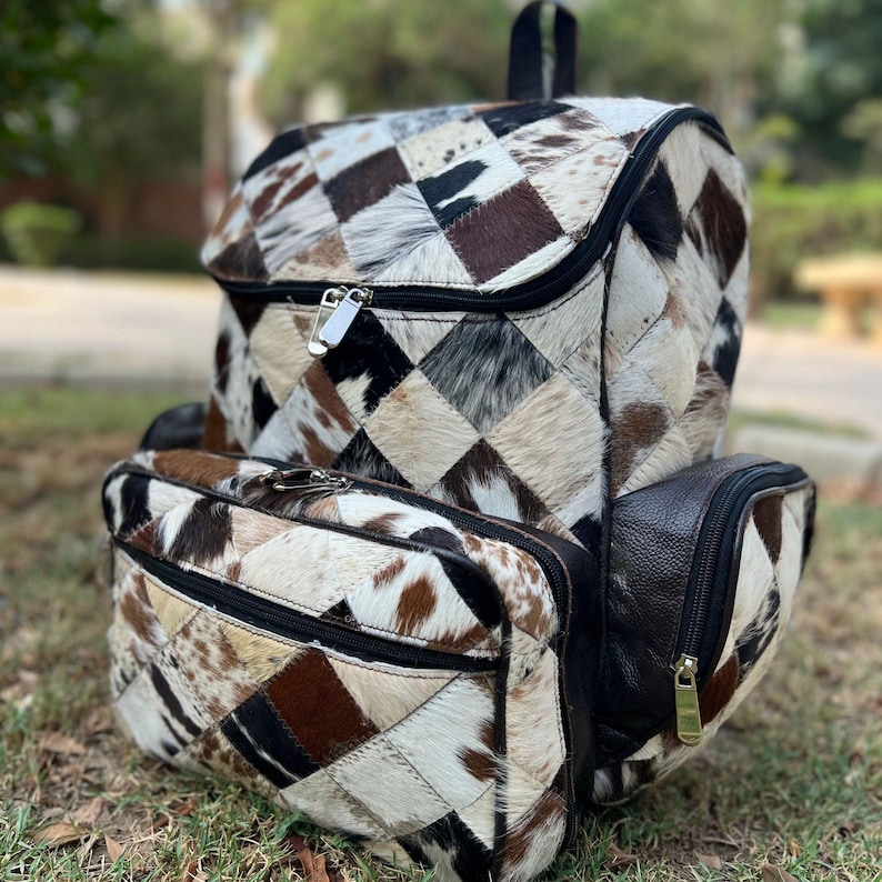 May include: A patchwork backpack and matching pouch made from cowhide. The backpack features a flap closure, a zippered main compartment, and a front zippered pocket. The cowhide has a mix of brown, black, and cream-colored patches.