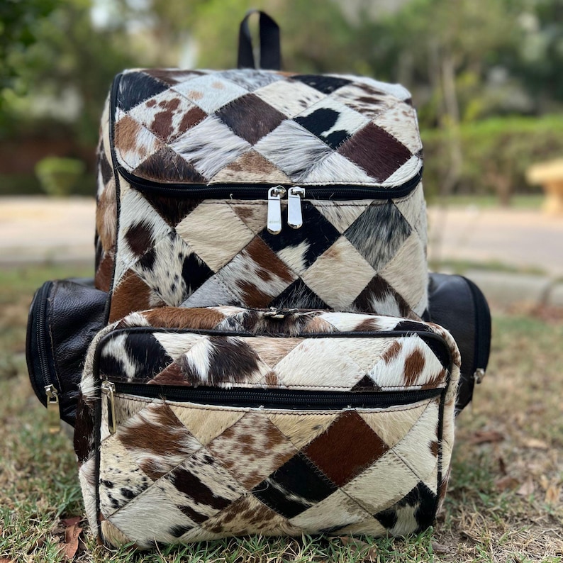 May include: A patchwork backpack with a front pouch, featuring a geometric pattern of brown, black, and cream-colored cowhide squares. The backpack has black leather accents and silver zippers.
