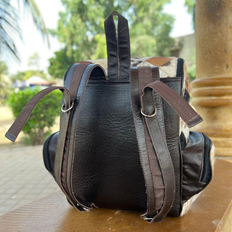 May include: A brown leather backpack with adjustable straps and a top handle. The backpack features a patterned flap with tan, brown, and cream colors. The bag has a front pocket and silver metal hardware.