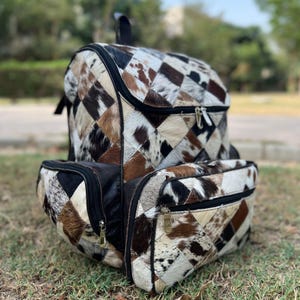 May include: A patchwork backpack with a geometric pattern of brown, black, and white cowhide. The backpack has a top handle, two zippered compartments, and two front pockets. The zippers and hardware are gold-toned.