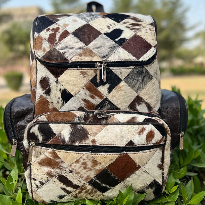 May include: A multi-toned cowhide backpack with a flap closure and a front zippered pocket. The backpack features a patchwork design of brown, black, and cream-colored squares. The bag has a top handle and adjustable straps.