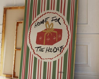 May include: A framed holiday art print with a red gift box and the text "HOME FOR THE HOLIDAYS" on a striped background of red, green, and white. The artwork is leaning against a white wall and wooden floor.