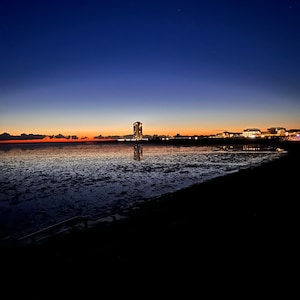 May include: A dark silhouette of a city skyline with a tall building lit up against a deep blue sky with a few stars visible. The city is reflected in the calm water of the ocean.