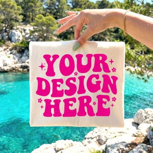以下が含まれることがあります： 鮮やかなピンク色の「YOUR DESIGN HERE」の文字がレトロなスタイルでプリントされた、クリーム色のトートバッグ。 ターコイズブルーの海と岩の多い海岸線を背景に、バッグが掲げられています。