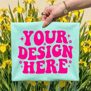 May include: A light blue t-shirt with the text "YOUR DESIGN HERE" in bright pink. The text is surrounded by pink stars and flowers. The shirt is held up in front of yellow flowers.