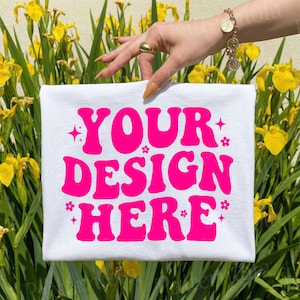 May include: White t-shirt with the text "YOUR DESIGN HERE" in bright pink, surrounded by stars and flowers. The shirt is held up against a backdrop of yellow flowers and green foliage.