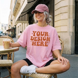 Puede incluir: Una camiseta rosa claro con el texto "YOUR DESIGN HERE" en naranja. La persona lleva una gorra rosa, pantalones cortos negros y zapatillas blancas. Están sentados en una mesa con una bebida.