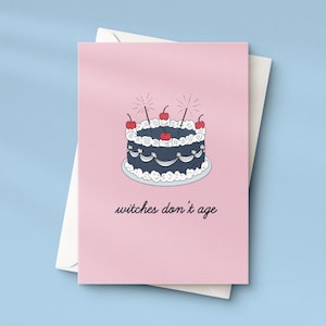 Könnte beinhalten: Rosa Grußkarte mit einer Kuchenillustration und dem Text "witches don't age". Der Kuchen ist dunkelblau mit weißem Zuckerguss und roten Kirschen. Die Karte liegt auf einem weißen Umschlag, vor hellblauem Hintergrund.