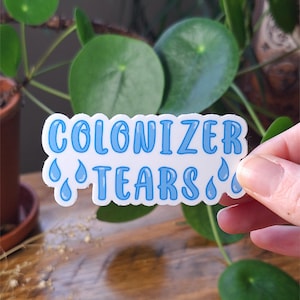 Könnte beinhalten: Ein gestanzter Aufkleber mit weißem Hintergrund, der den Text COLONIZER TEARS in einer fetten, abgerundeten, hellblauen Schrift zeigt. Das Design enthält mehrere hellblaue Tränenformen, die um den Schriftzug herum angeordnet sind.