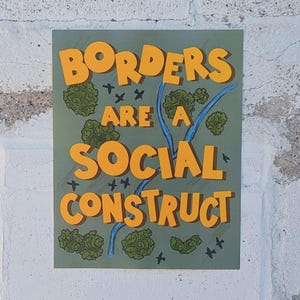 May include: A rectangular art print with the words "BORDERS ARE A SOCIAL CONSTRUCT" in orange block letters. The background is green with blue lines and green circular shapes. The print is mounted on a white brick wall.