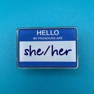 May include: Blue plastic name tag with the text "Hello, my pronouns are she/her".