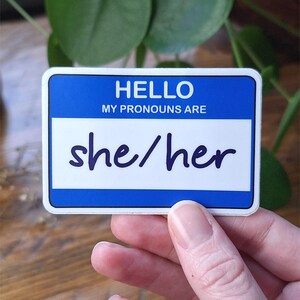 May include: A rectangular sticker designed like a name tag with a blue border and white centre. The text HELLO MY PRONOUNS ARE is printed in white at the top, and the handwritten-style text she/her is printed in dark blue in the centre section.