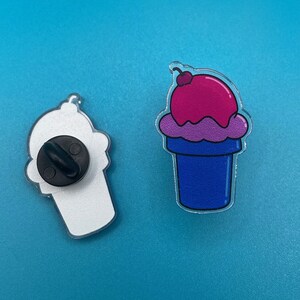 May include: Two ice cream cone pins, one white and one with pink and blue ice cream. The white cone has a black pin back. The blue cone has a red cherry on top.