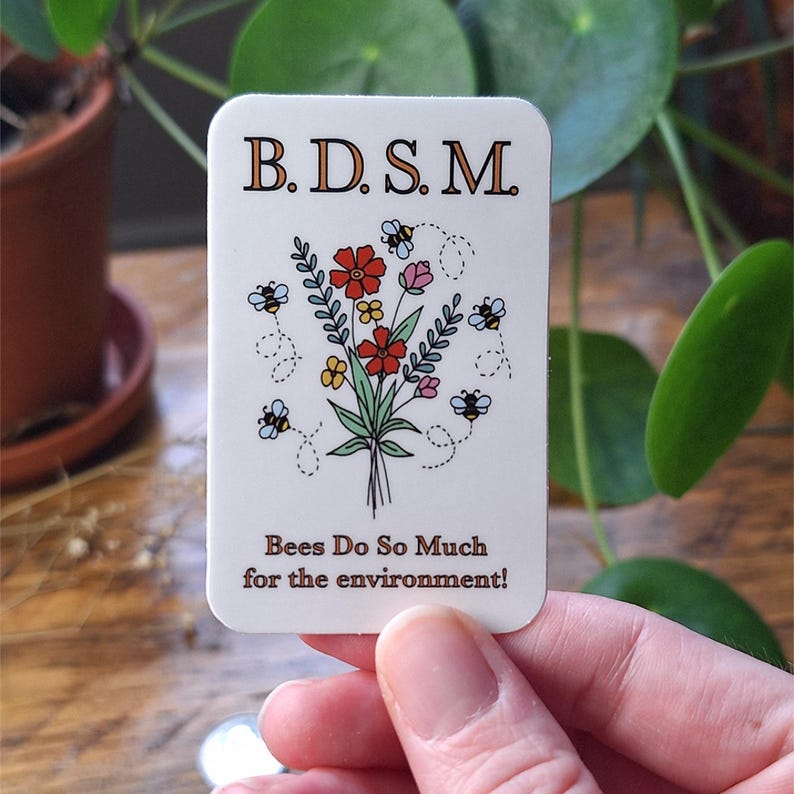 May include: A rectangular white sticker featuring the text B.D.S.M. at the top and Bees Do So Much for the environment! at the bottom. The center displays a colorful illustration of a wildflower bouquet surrounded by four small flying bees.