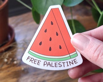 May include: A triangular sticker shaped like a watermelon slice with a red interior, black seeds, and a light green rind. The white base of the sticker features the text "FREE PALESTINE" printed in dark brown capital letters.