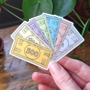 May include: A fan of five rectangular stickers designed to look like colorful play money. Each bill features the text EAT THE RICH and a specific denomination value of 500, 100, 50, 20, 10, or 5 in yellow, purple, green, blue, and pink colors.