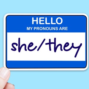 May include: A rectangular sticker designed like a name tag with a blue border and white centre. The text HELLO MY PRONOUNS ARE is printed in white at the top. The handwritten text she/they is written in dark blue ink across the white centre section.